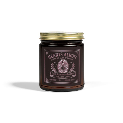 A circular, dark amber glass jar candle with a solid gold-colored metallic lid. The glass jar has a large, rectangular black label with a light-colored decorative border and ornate scrollwork. The label features the title "Hearts Alight" at the top in elegant script and lists the scent notes "Exotic Cardamom, Coconut, and Zesty Lemon" below it. A single white cotton wick with a lit flame is visible, centered within the wax. The wax itself is a rich, warm, orange-amber color, consistent with the jar's hue.
